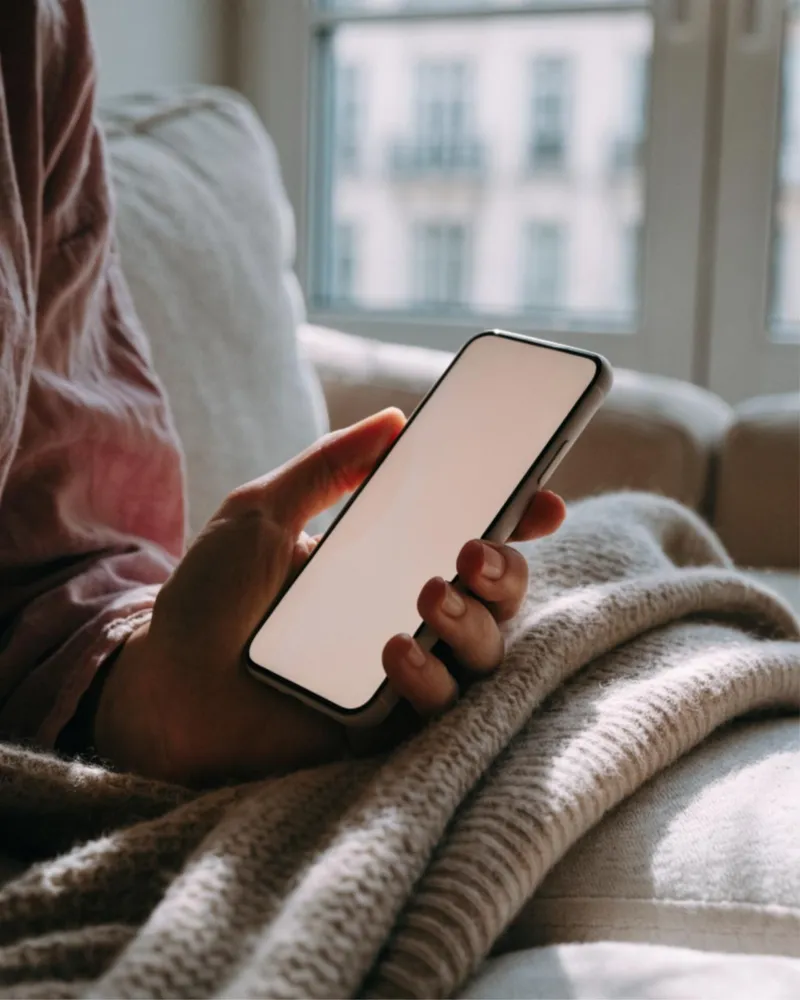 ソファでブランケットをかけ、スマートフォンを手に持つ人の手元